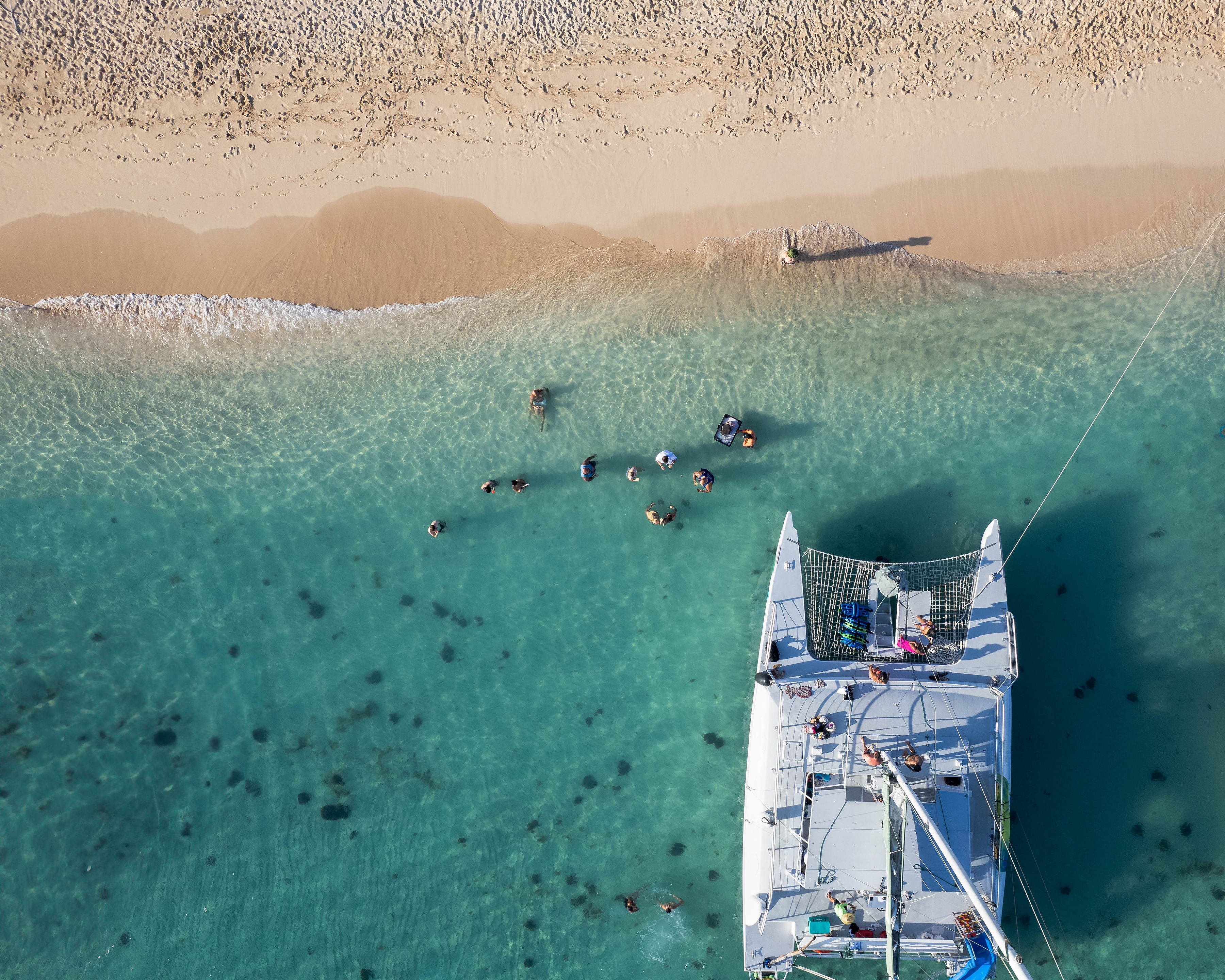 Aerial view of a catamaran near a beach with people swimming in clear water.